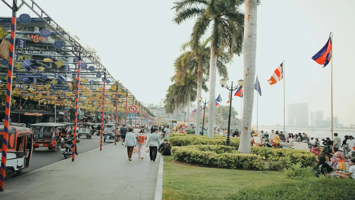 Bustling scene along Phnom Penh riverside with tuk-tuks, people, and flags creating a lively atmosphere.