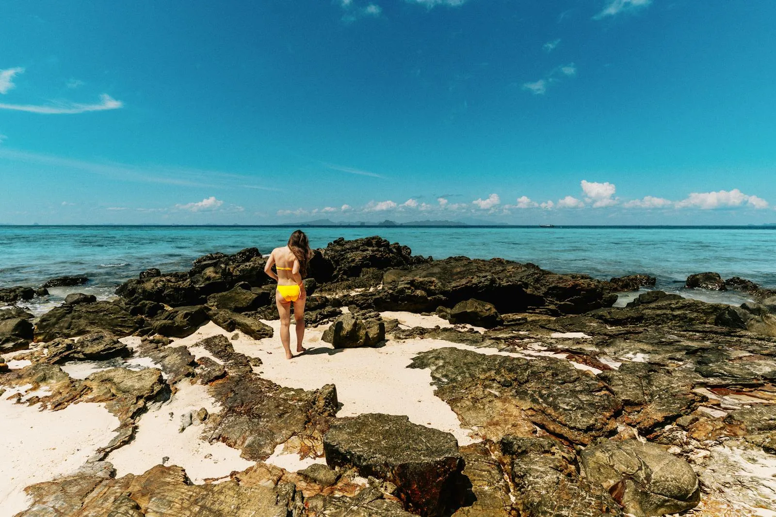 Woman in yellow bikini exploring rocky beach in Krabi, Thailand under a bright blue sky.